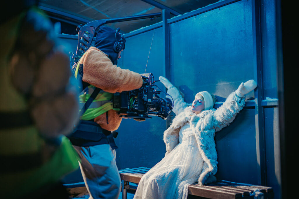 Camera crew filming Monetochka at a blue bus stop on the Self-Harm music video set